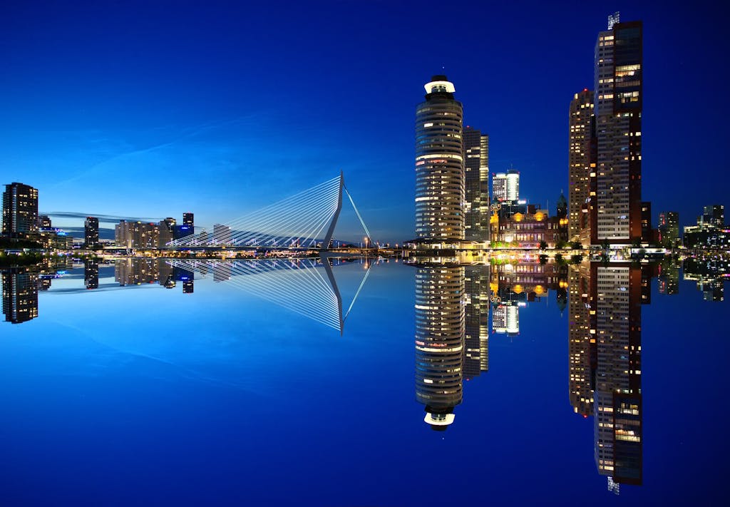Beautiful night view of Rotterdam skyline with illuminated buildings and Erasmus Bridge reflection.